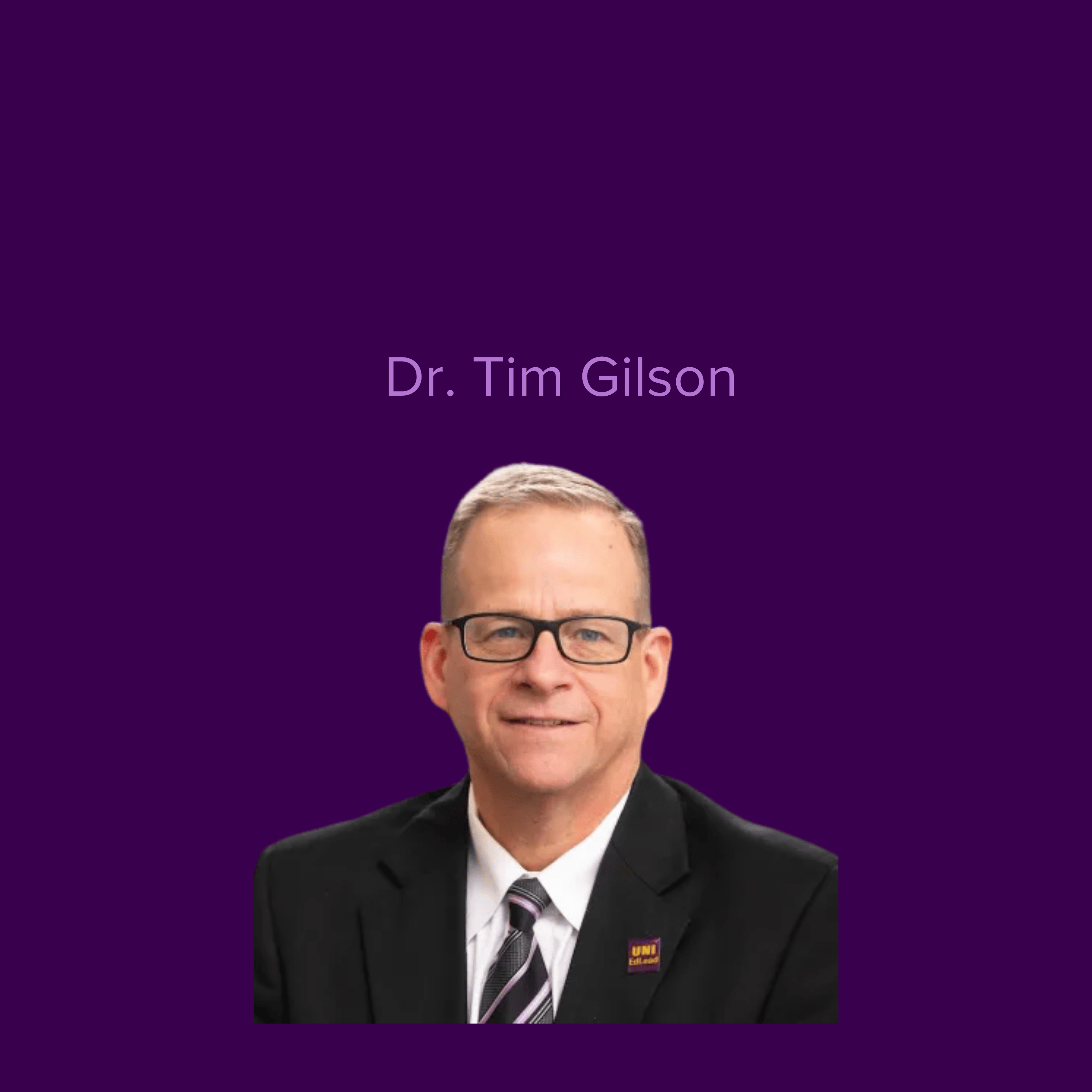 Image of Tim Gilson wearing glasses and a dark suit with a UNI button
